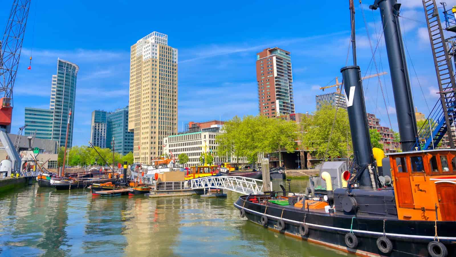 Boats docked on a river with modern skyscrapers and green trees in the background under a blue sky.