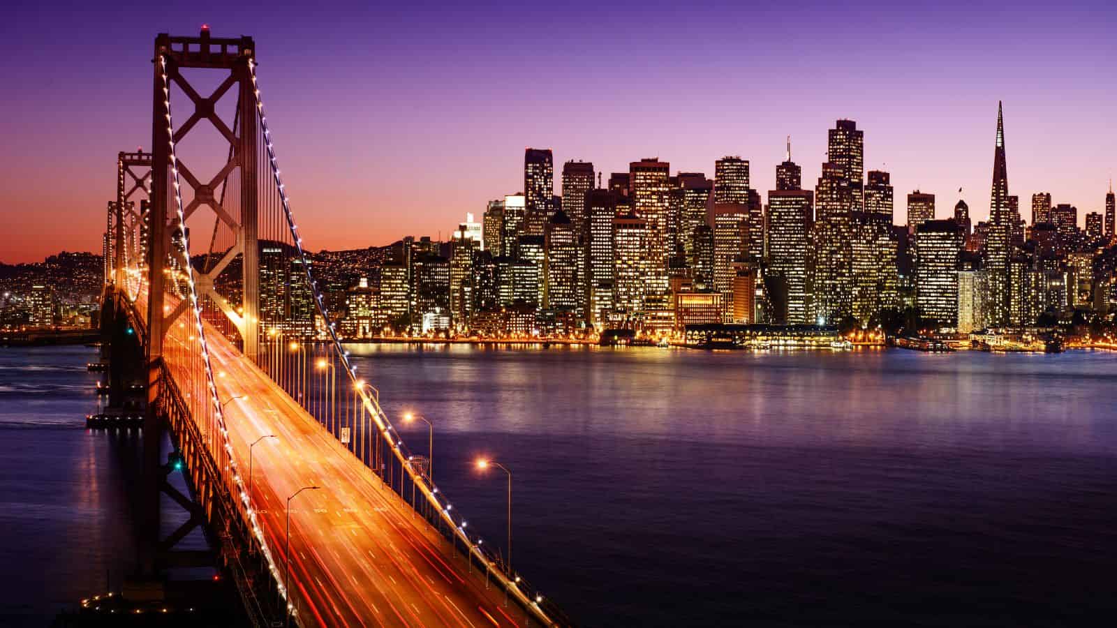 San Francisco Bay Bridge at dusk with city skyline illuminated against a purple and orange sky.
