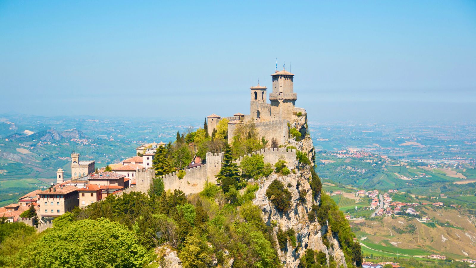 Ancient fortress towers atop a rocky hill, overlooking a lush green valley and distant landscape under a clear sky.