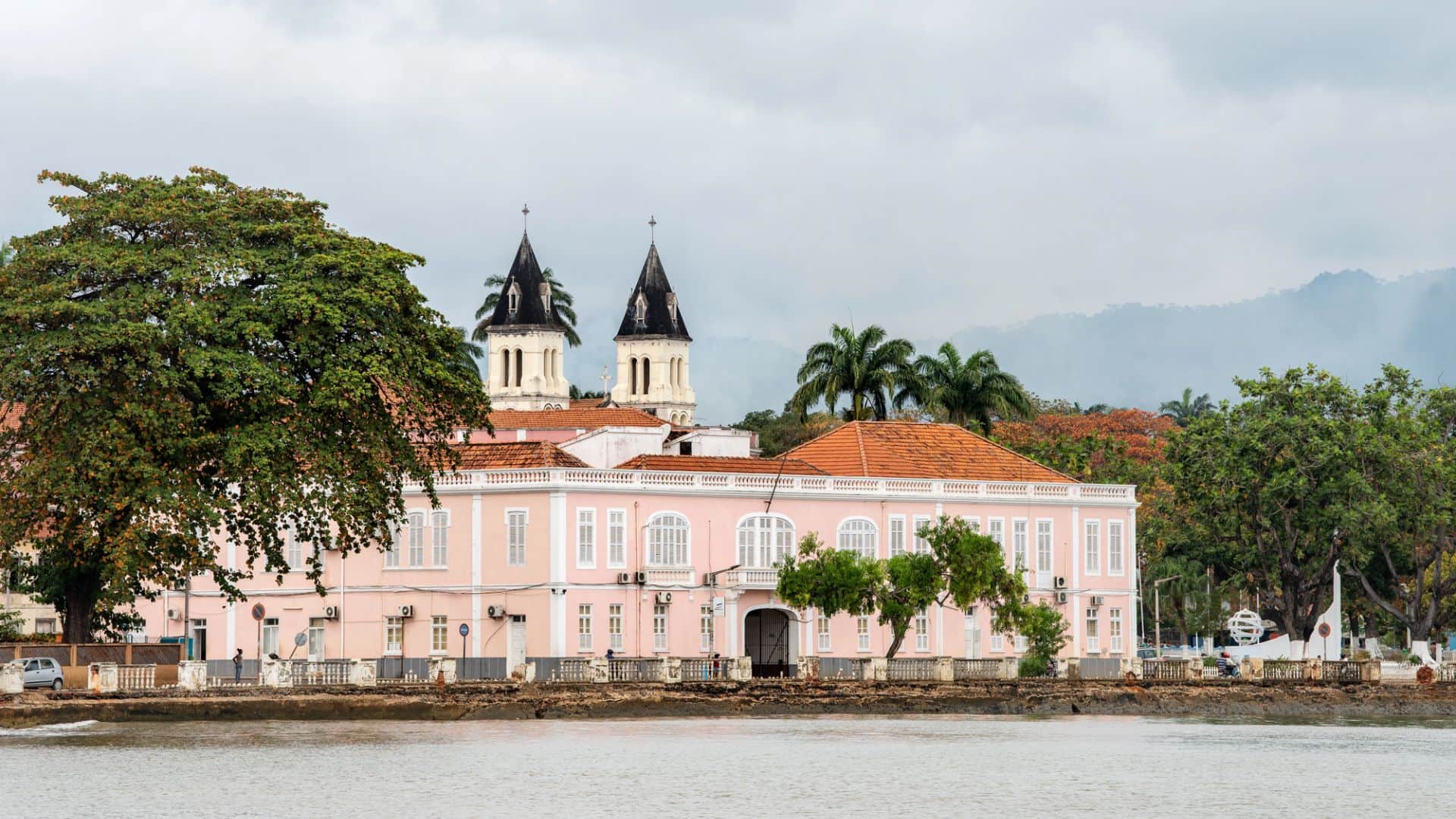Pink colonial-style building with twin church towers by the water, surrounded by trees under a cloudy sky.