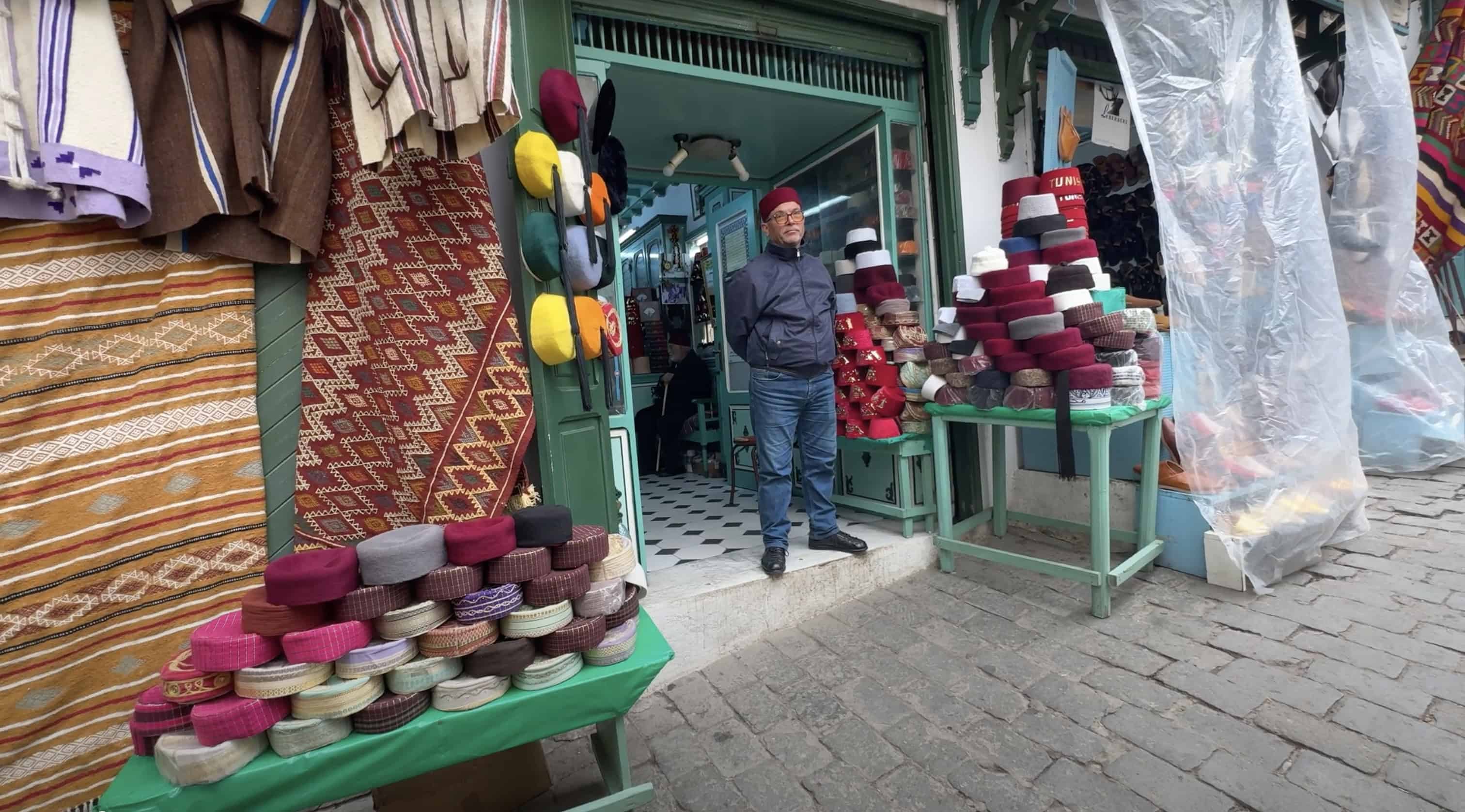 A man stands at a shop entrance selling colorful hats, with hats displayed on tables outside—a lively scene you might spot during your Tunisia itinerary 1 day adventure.