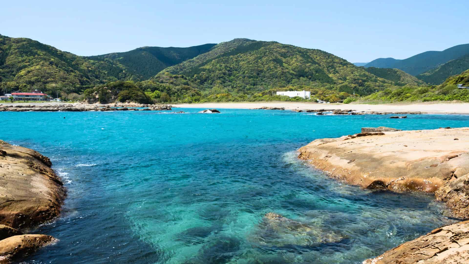 Clear blue water with rocky shores and green hills in the background under a bright sky.