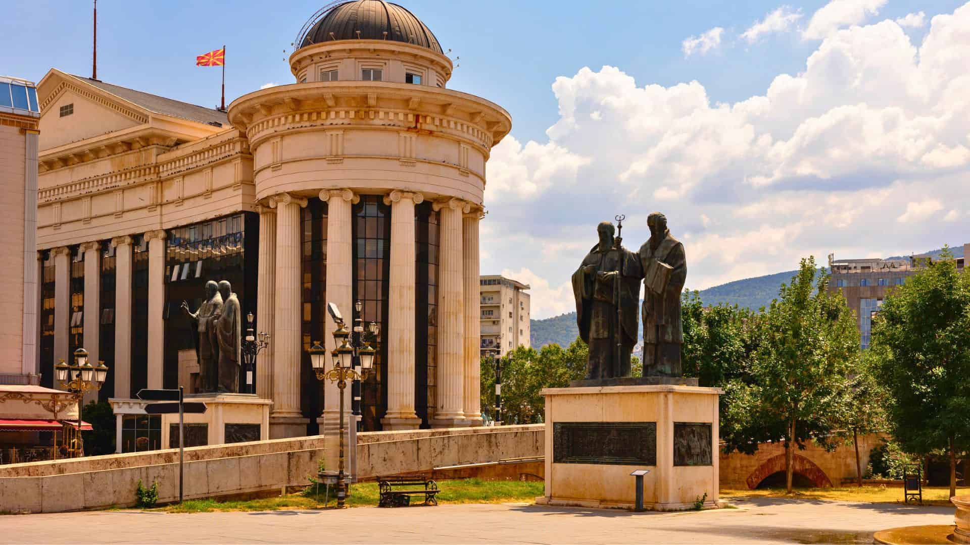 Statues in front of a neoclassical building with columns in a sunny city square.