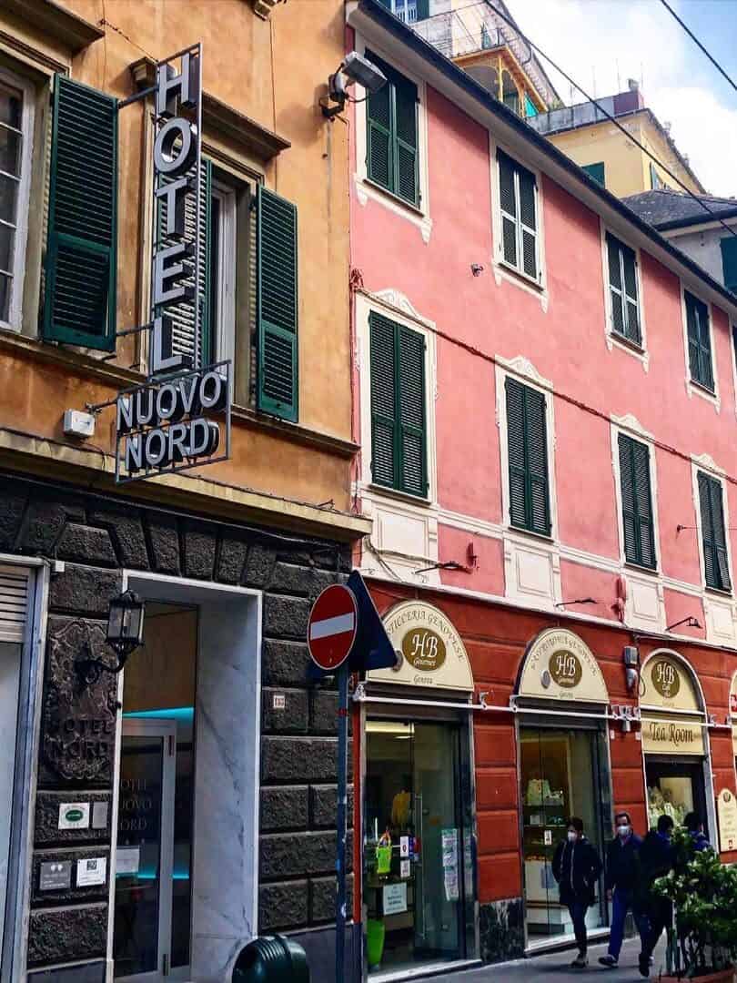 People walk past colorful buildings and shops with a “HOTEL NUOVO NORD” sign in a lively Genoa street scene, capturing the vibrant atmosphere of Italy.