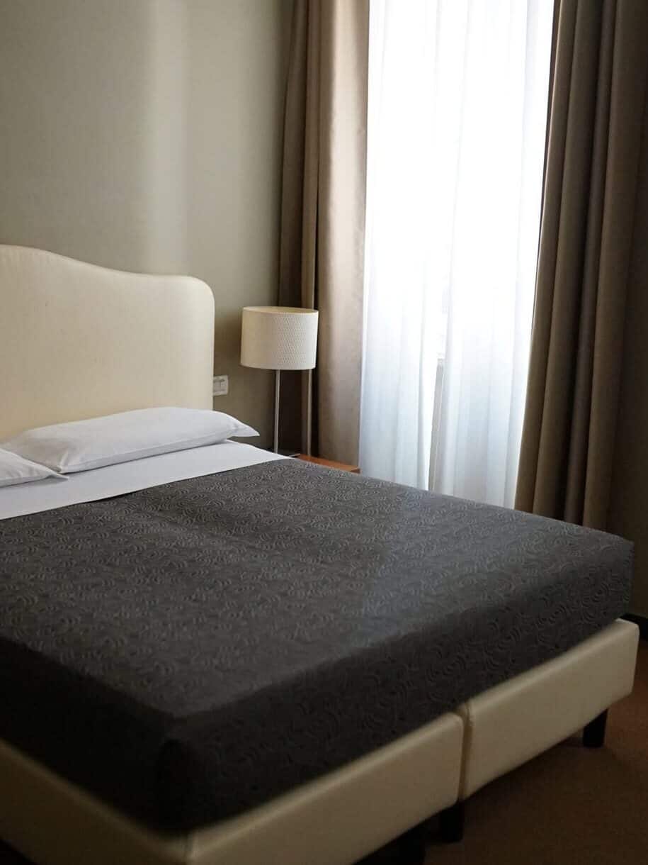 A neatly made bed with gray bedding sits beside a lamp and window with sheer curtains in a simple room—perfect for unwinding after 24 hours in Genoa, Italy.
