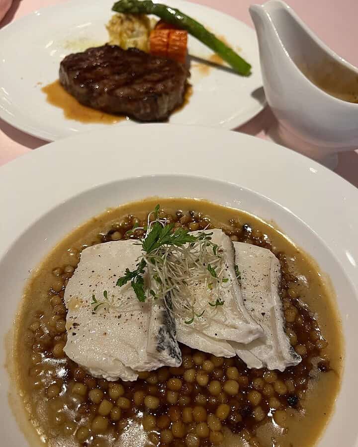 A plate of fish on couscous with sauce, showcasing the elegance of cruise ship food, with a steak and vegetables in the background.