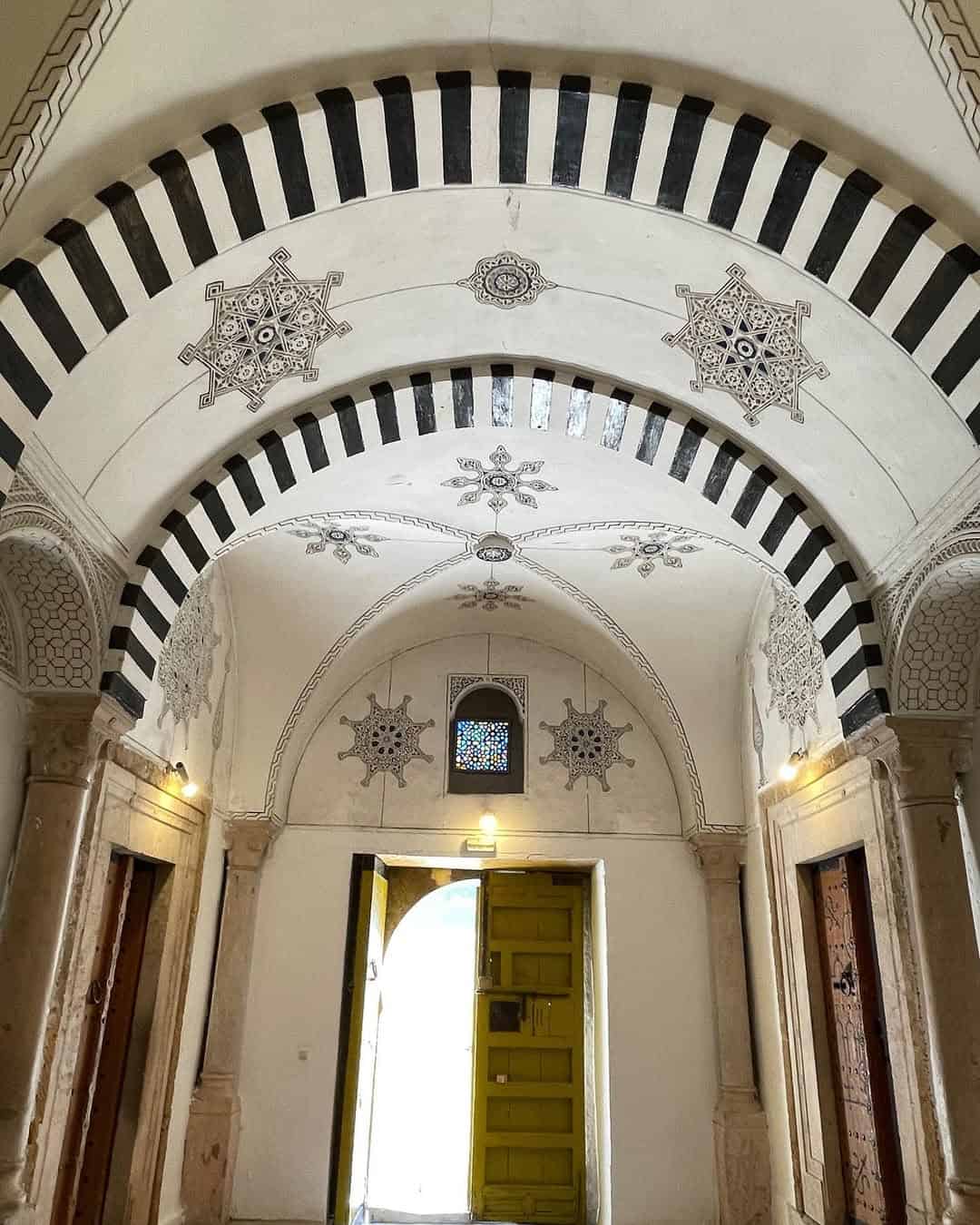 Arched hallway with ornate black-and-white ceiling patterns, reminiscent of a Tunisia itinerary 1 day adventure, and an open yellow door letting in sunlight.