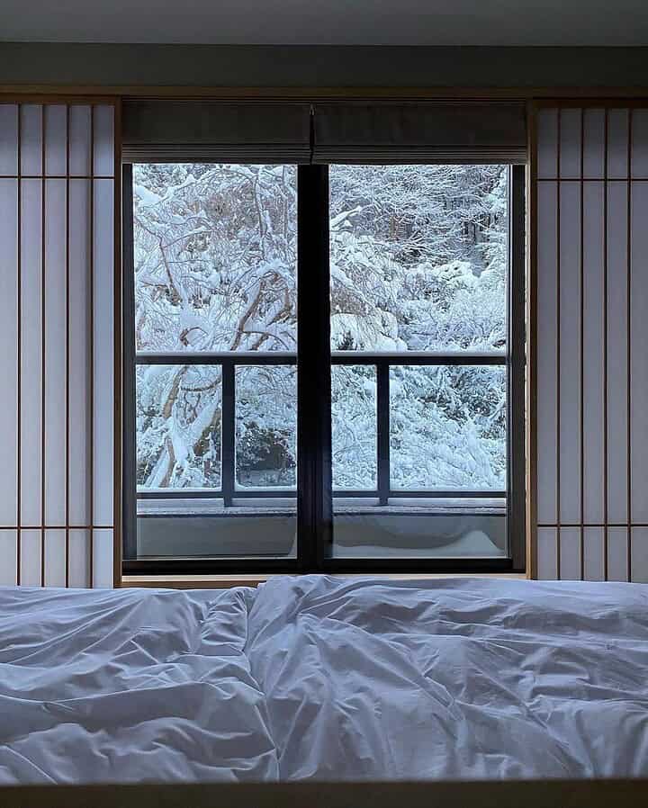 Unmade bed in front of a window overlooking a snowy landscape and trees, capturing the cozy allure of winter travel.