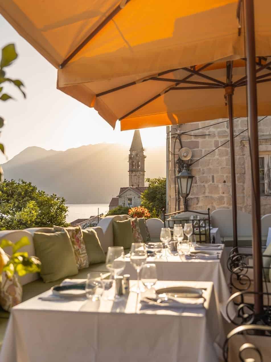 Outdoor restaurant with set tables under umbrellas, overlooking a tower and mountains at sunset—a perfect spot to savor Italy’s charm during 24 hours in Genoa.