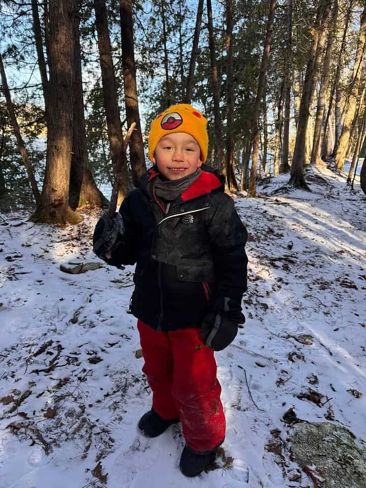 Smiling child in a yellow hat and red pants stands in a snowy, wooded area holding a stick, enjoying winter travel adventures.