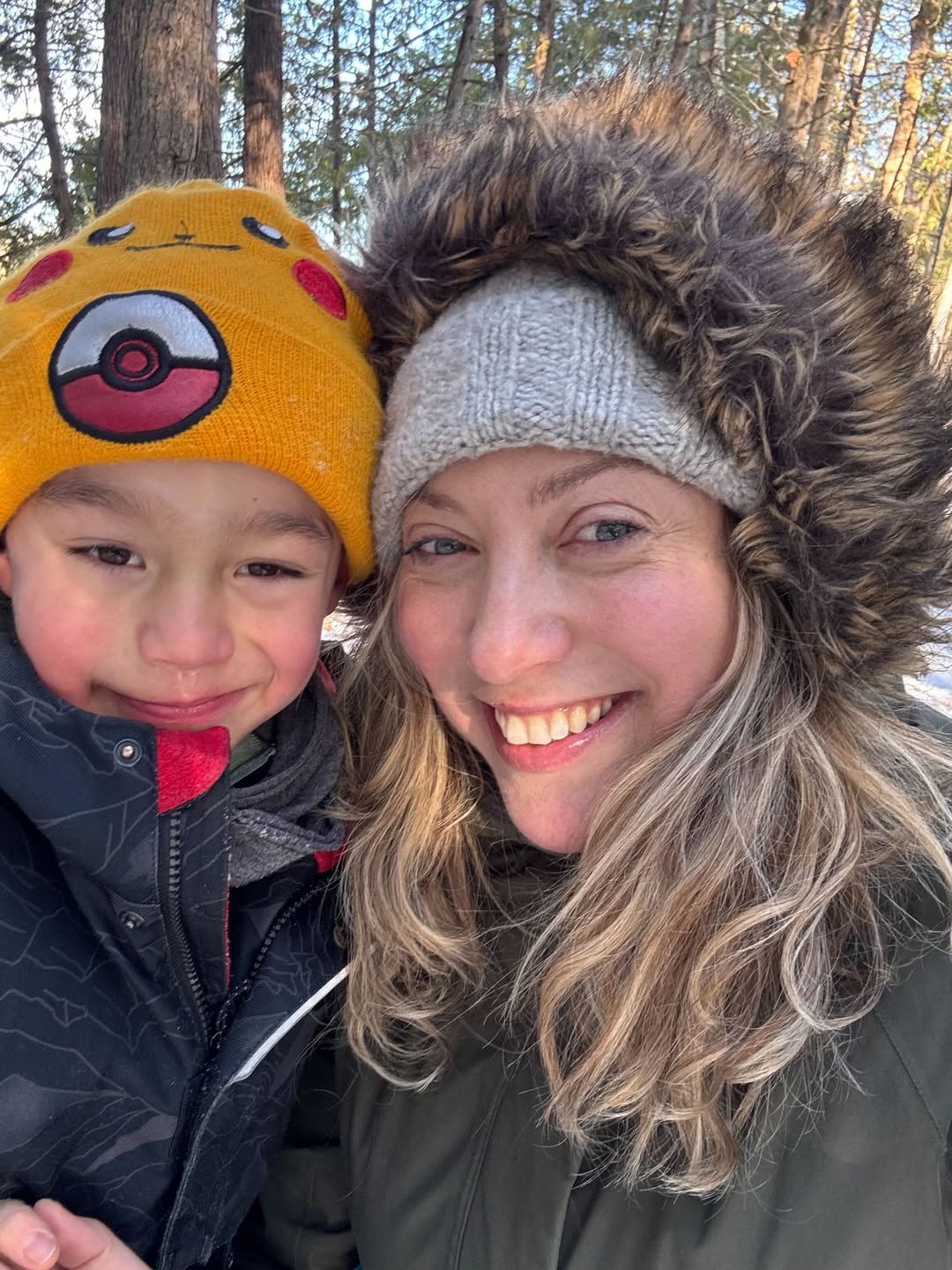 Woman and child bundled in winter coats, smiling outdoors with trees in the background, enjoying the magic of winter travel together.