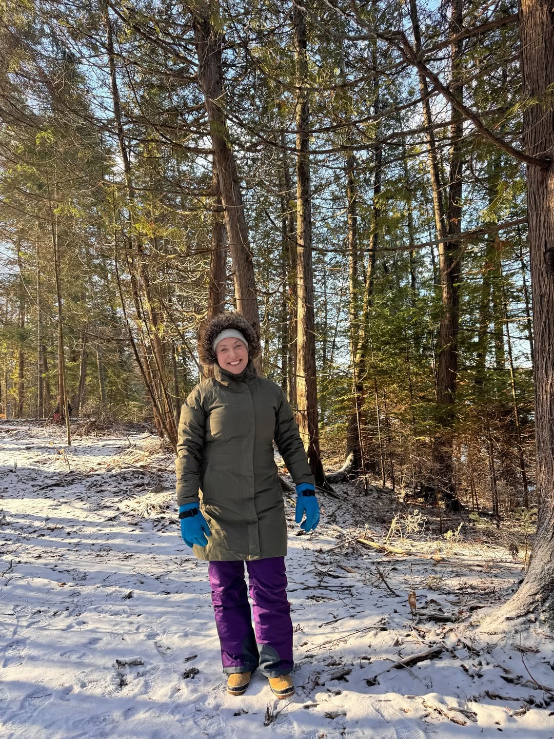 A person in winter clothes stands on a snowy forest path with sunlight streaming through the trees, capturing the magic of winter travel.