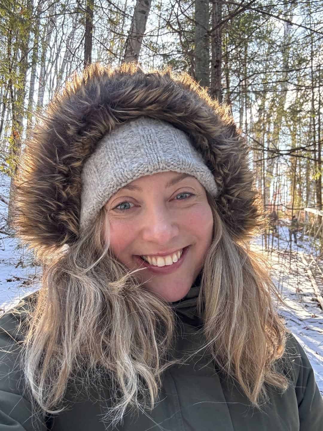 Woman in a winter coat with fur-lined hood smiles outside in a snowy, sunlit forest, embracing the magic of winter travel.