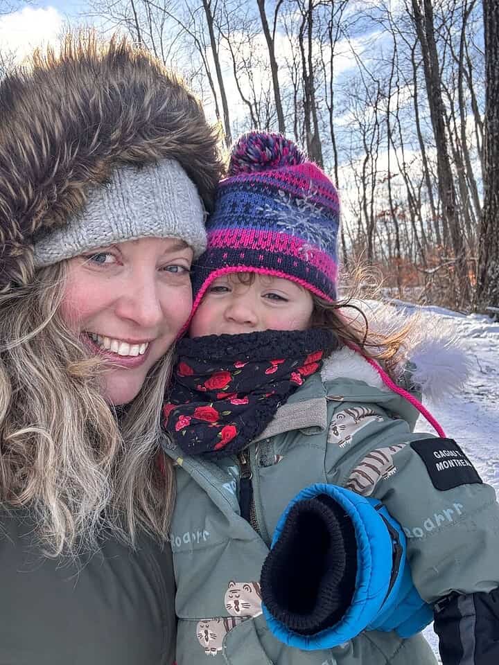 Woman and young child in winter coats smiling outdoors, surrounded by snowy trees and bright sunlight&mdash;capturing the joy of winter travel together.