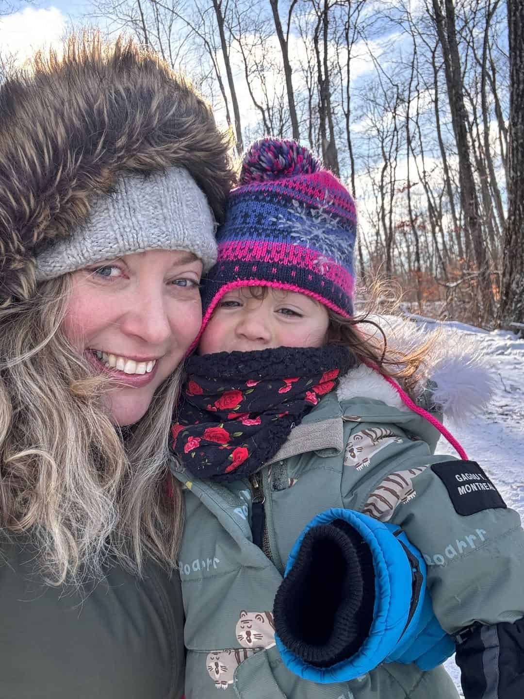 Woman and young child in winter coats smiling outdoors, surrounded by snowy trees and bright sunlight—capturing the joy of winter travel together.