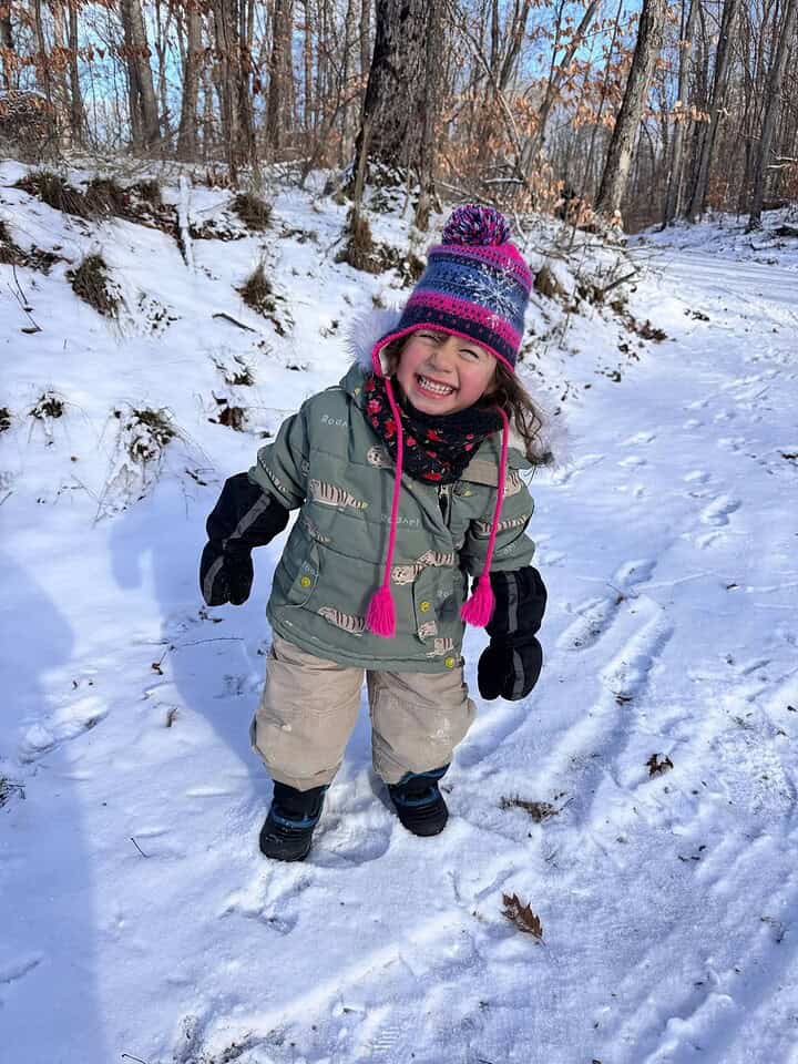 Smiling child in winter clothes stands on a snowy forest path, enjoying the magic of winter travel with trees and blue sky in the background.