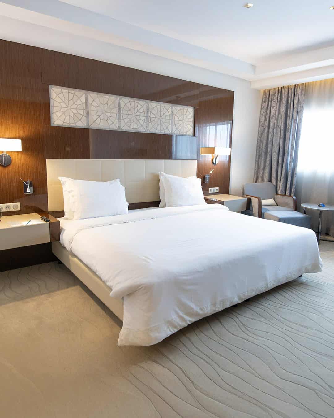 Modern hotel room with a large bed, white bedding, bedside tables, armchair, and natural light from a window—a perfect retreat after exploring your Tunisia itinerary 1 day adventure.