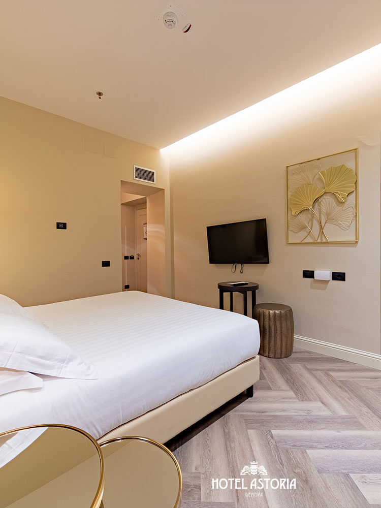 Modern hotel room in Genoa with a comfortable bed, wall-mounted TV, and a view into an adjoining room with another bed—ideal for your stay in Italy, whether you’re here for 24 hours or a longer visit.