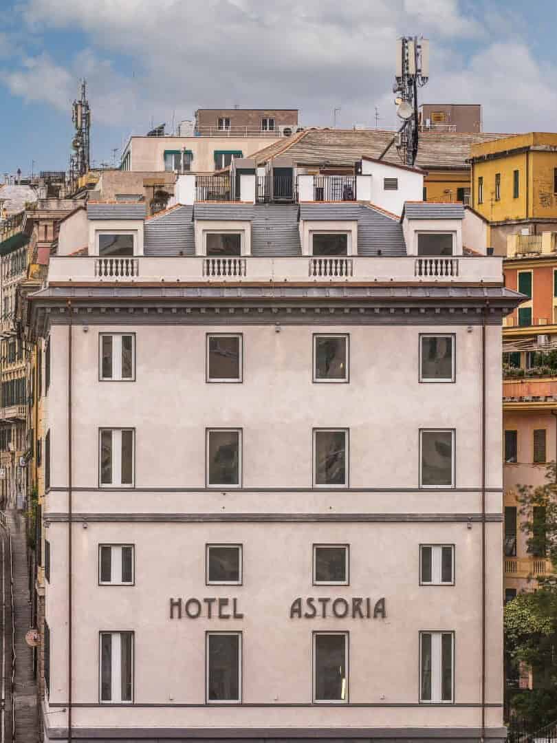 Hotel Astoria building with multiple windows, surrounded by colorful city buildings in Genoa, Italy, and a cloudy sky above.