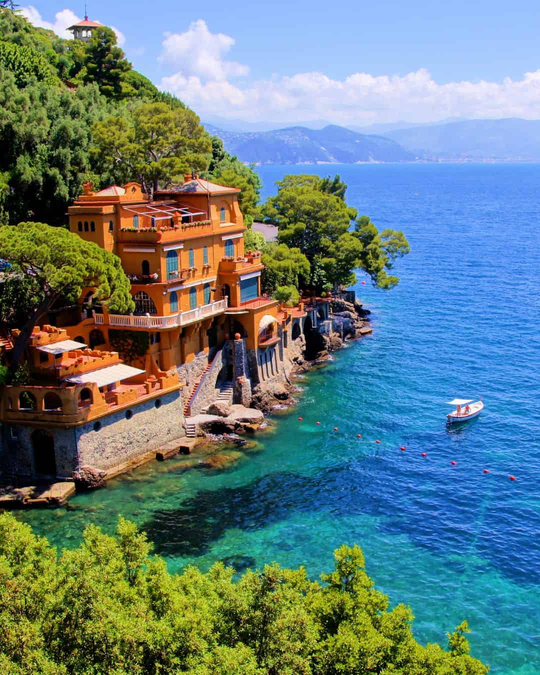 Colorful villa on the rocky coastline of Genoa, Italy, with lush trees, overlooking the clear blue sea and a small boat nearby—perfect for exploring in 24 hours.
