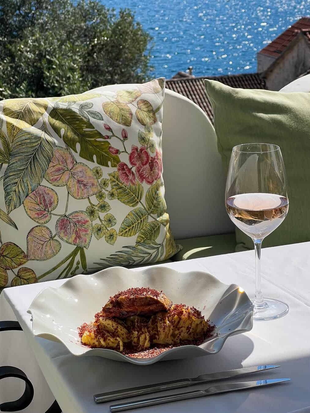 Enjoy a plate of food and a glass of rosé wine on a table with floral cushions, overlooking the stunning sea view—an ideal way to spend 24 hours in Genoa, Italy.