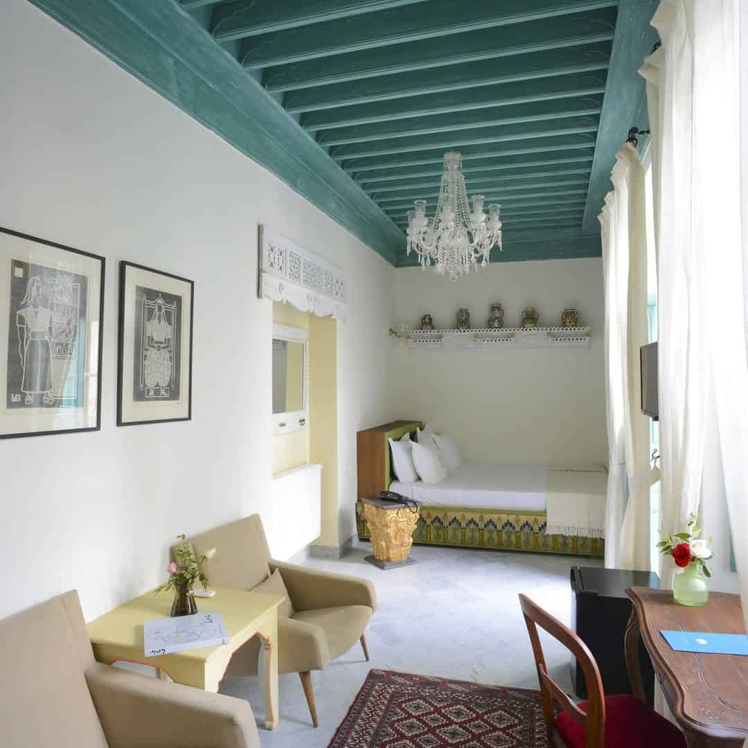 Bright room with teal ceiling, chandelier, cozy seating, bed, and decorative items on a shelf by large windows—perfect for planning your Tunisia itinerary 1 day adventure.