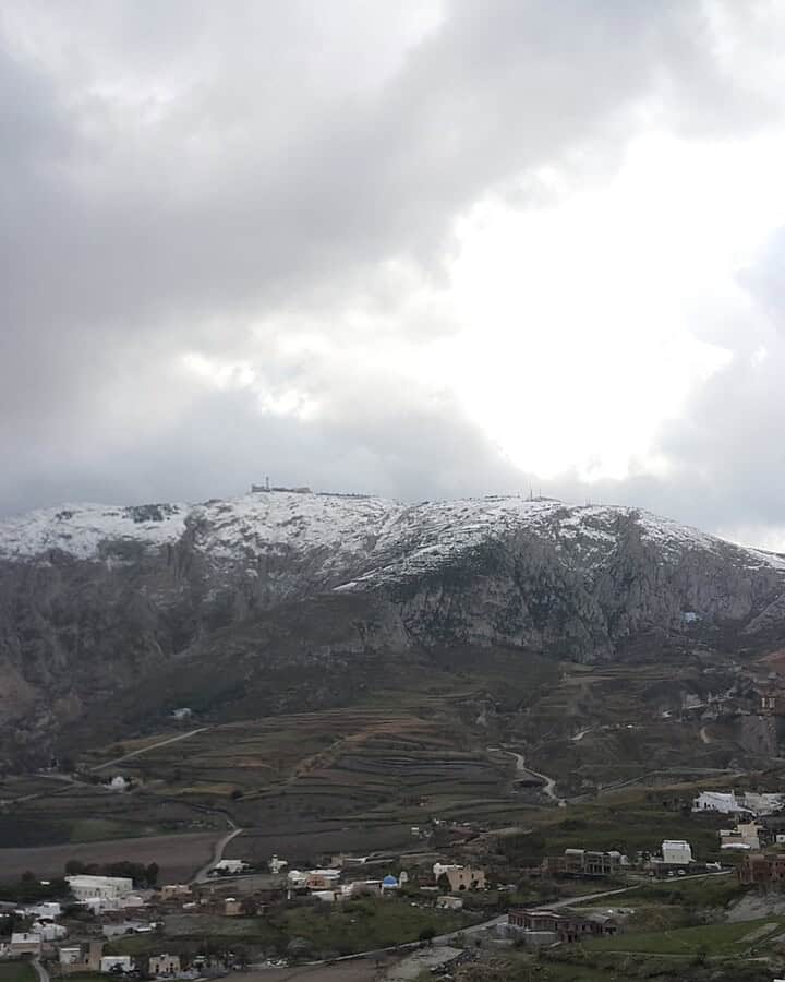 Snow-capped mountains under a cloudy sky set the scene for a picturesque village and terraced fields in the foreground&mdash;an ideal destination for winter travel.