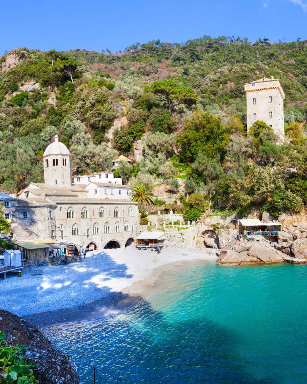 Stone abbey and tower by a pebbled beach with turquoise water, set against a green, wooded hillside—perfect for exploring during 24 hours in Genoa, Italy.
