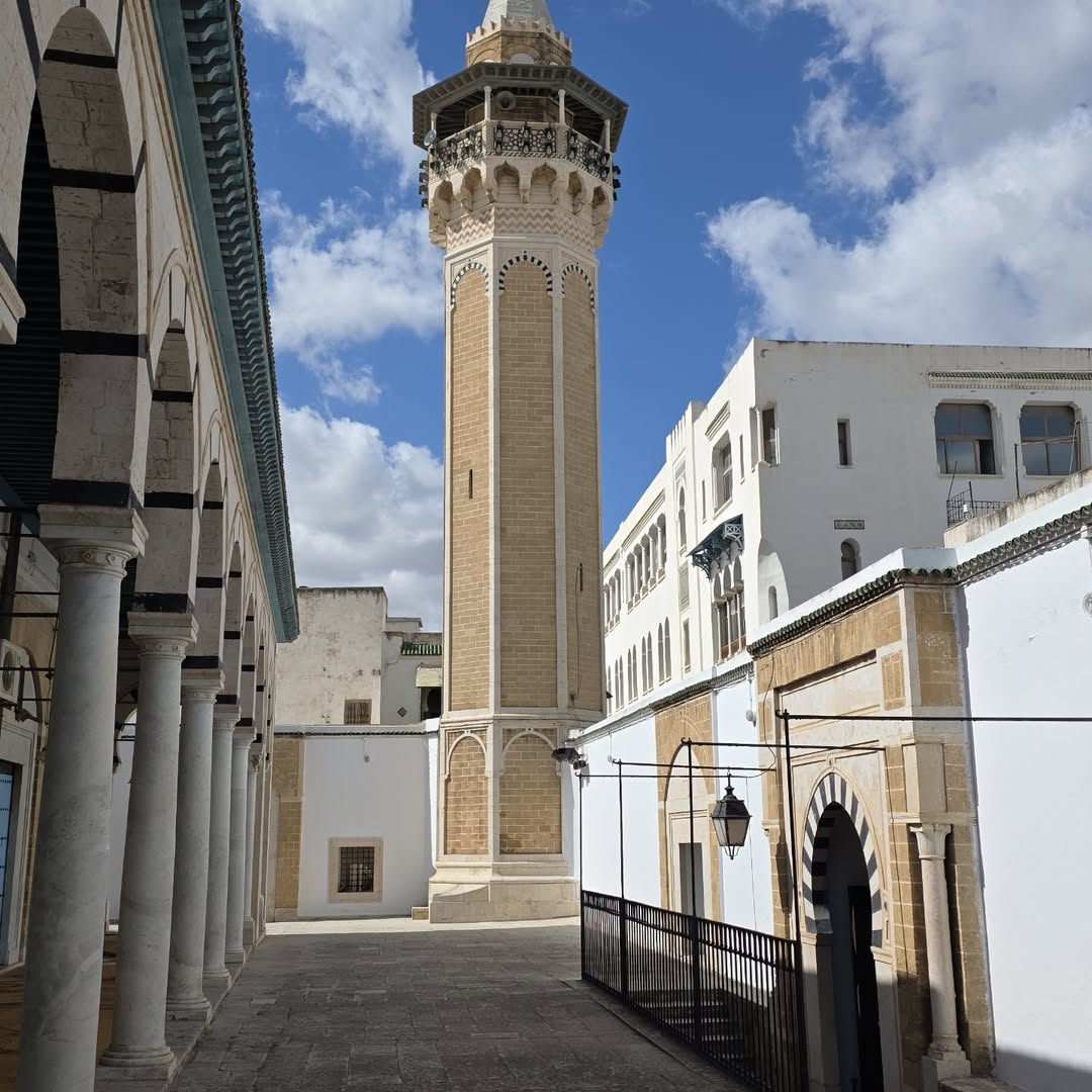 A tall minaret rises beside white buildings and arched colonnades under a cloudy blue sky—a must-see highlight on any Tunisia itinerary 1 day.