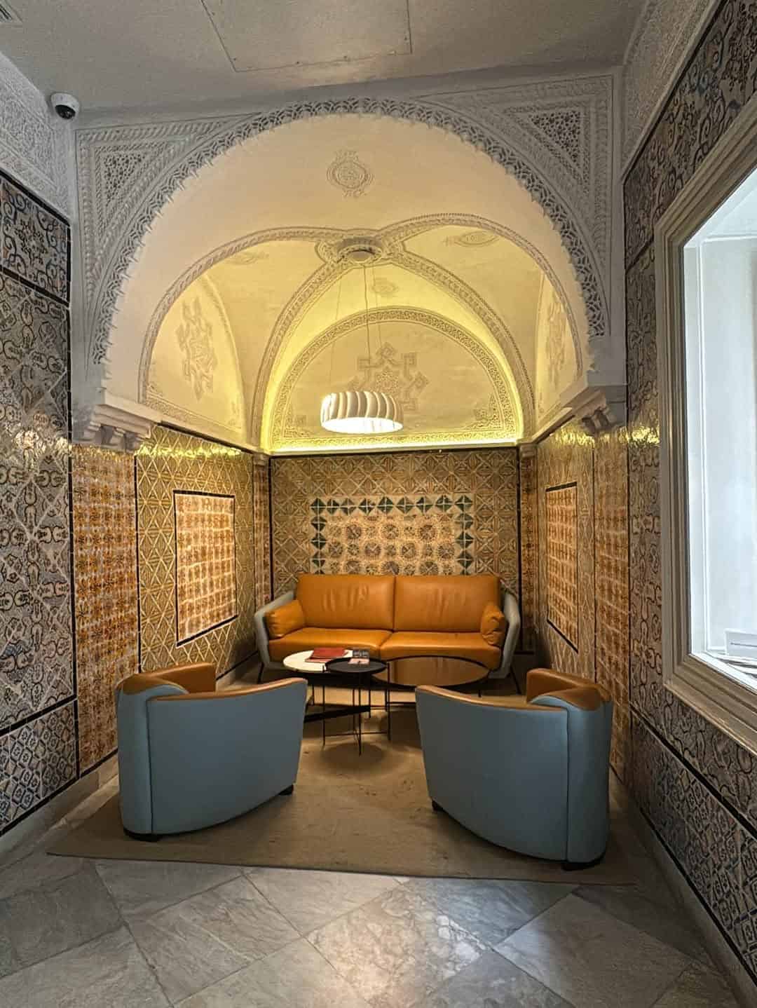 A cozy lounge with a tan couch, blue chairs, tiled walls, and an ornate arched ceiling—perfect inspiration for your Tunisia itinerary 1 day adventure.