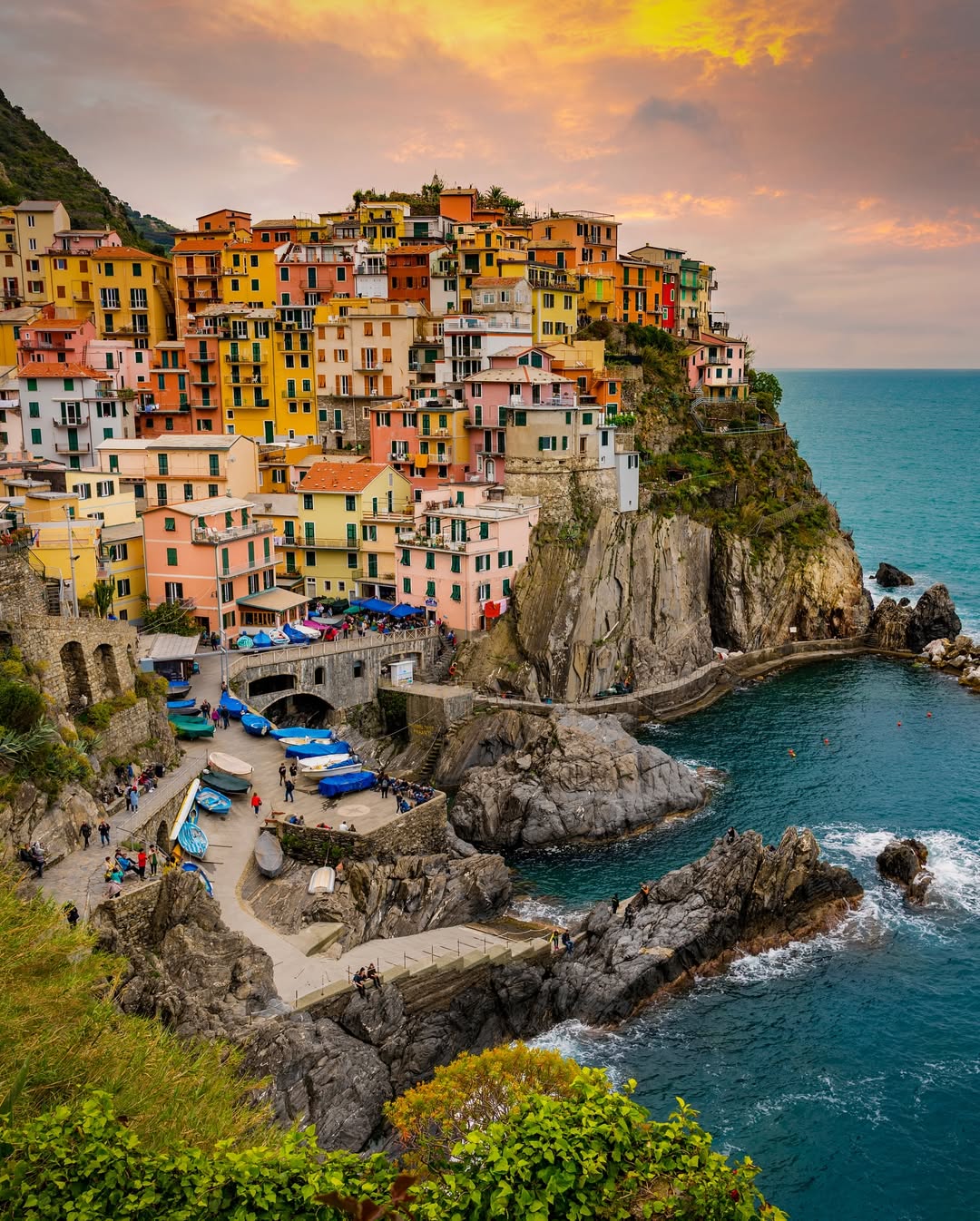 Colorful cliffside buildings in Genoa, Italy overlook a rocky coast and blue sea under a dramatic sunset sky—perfect for exploring during 24 hours in Genoa.