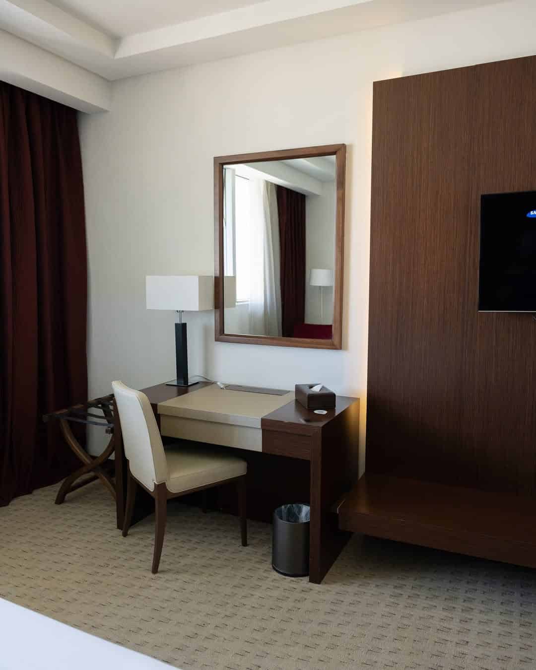 A hotel room desk with a chair, mirror, lamp, tissue box, and waste bin beside a TV on a wooden wall—ideal for planning your Tunisia itinerary 1 day adventure.