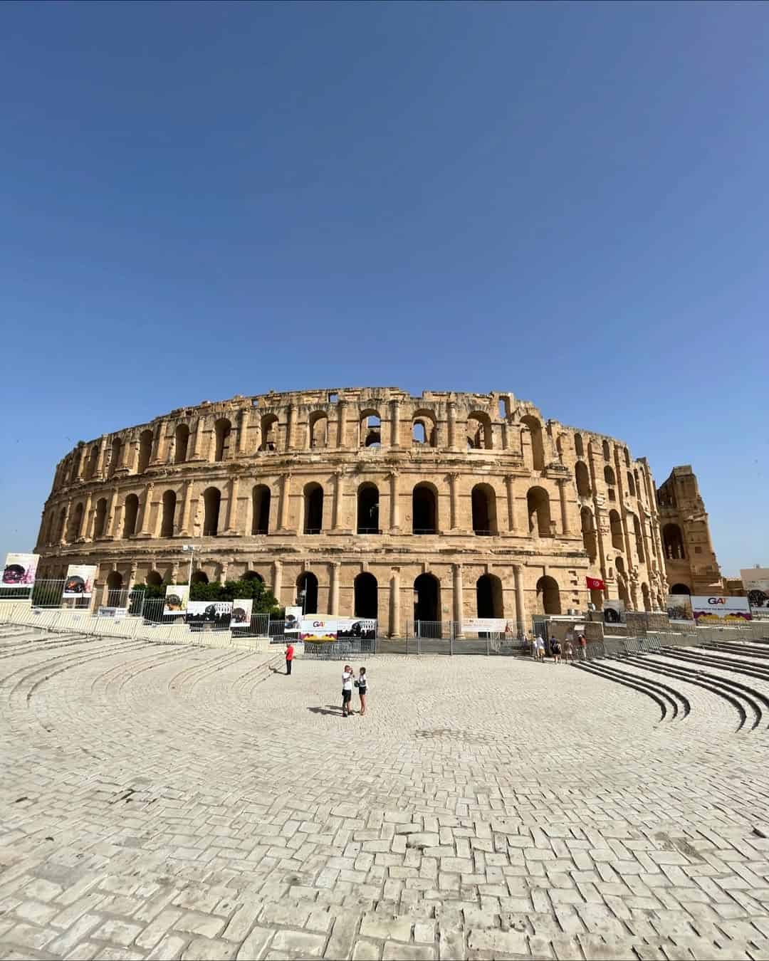 Ancient Roman amphitheater under a clear blue sky, with a few people standing in front on a stone courtyard—an essential stop on any Tunisia itinerary 1 day adventure.