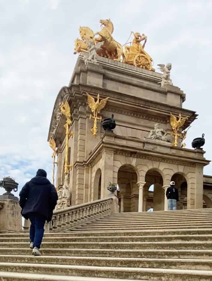A person climbs stairs toward a grand monument with golden statues and ornate architectural details, capturing the essence of a perfect Barcelona travel experience.