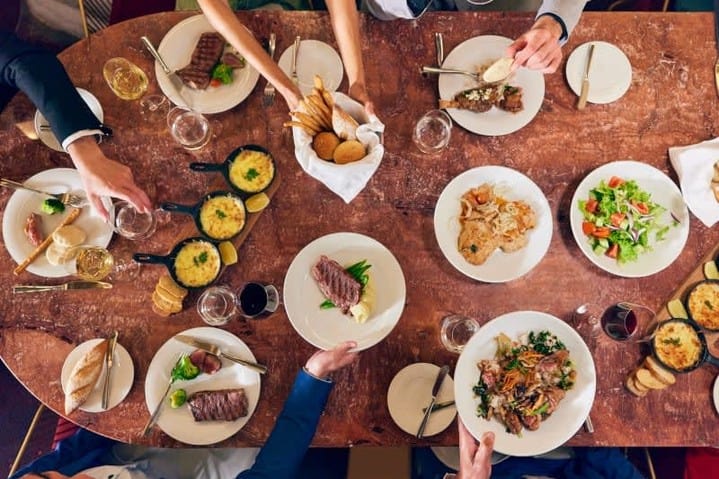 A group of people dining at a table with plates of steak, salad, bread, wine, and side dishes while enjoying exquisite cruise ship food during their voyage at sea.