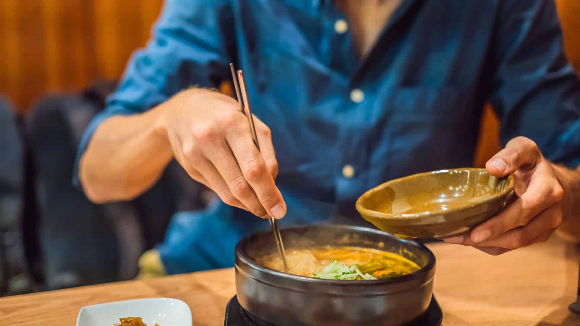 A person in a blue shirt eats hot soup with chopsticks and a small bowl at a wooden table.