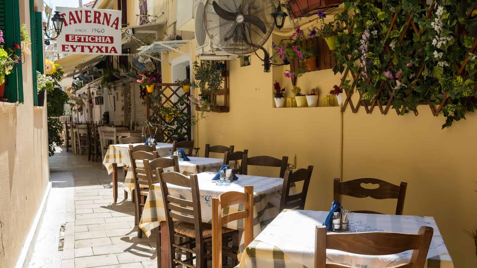 Outdoor tables and chairs line a narrow street next to a yellow wall with flowers and a “Taverna” sign above.