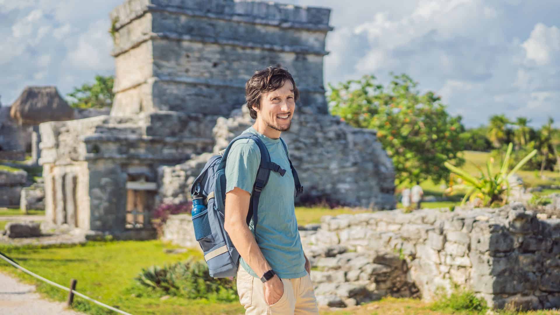 Man with a backpack smiling at ancient stone ruins on a sunny day with greenery around.