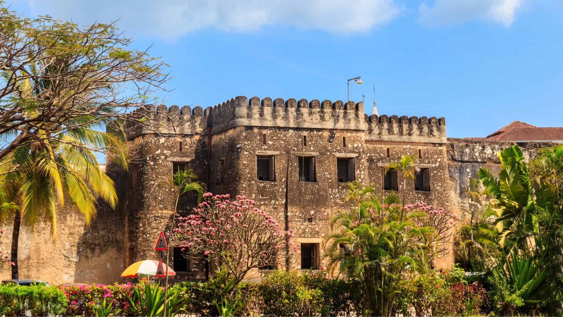 Old stone fort surrounded by lush green plants and flowering trees under a blue sky with some clouds.