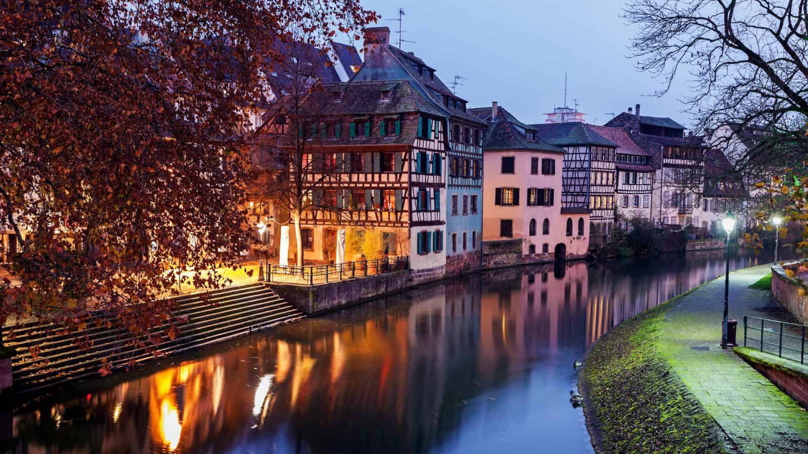 Half-timbered houses line a calm canal at dusk in Strasbourg, with reflections in the water.