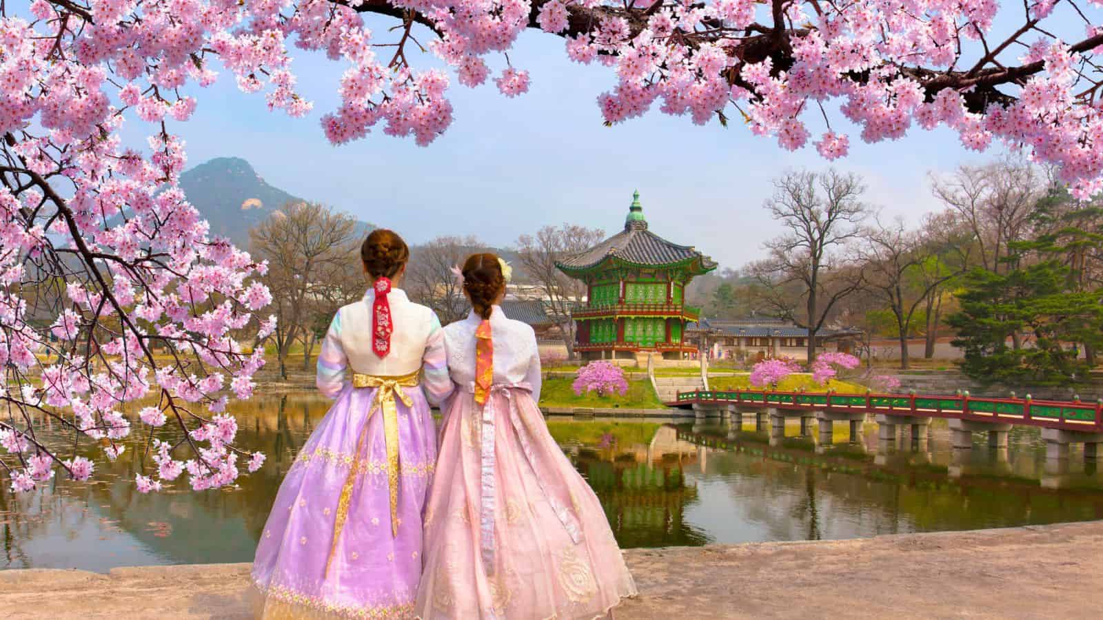 Two women in hanbok stand by a pond with cherry blossoms and a traditional Korean pavilion in the background.