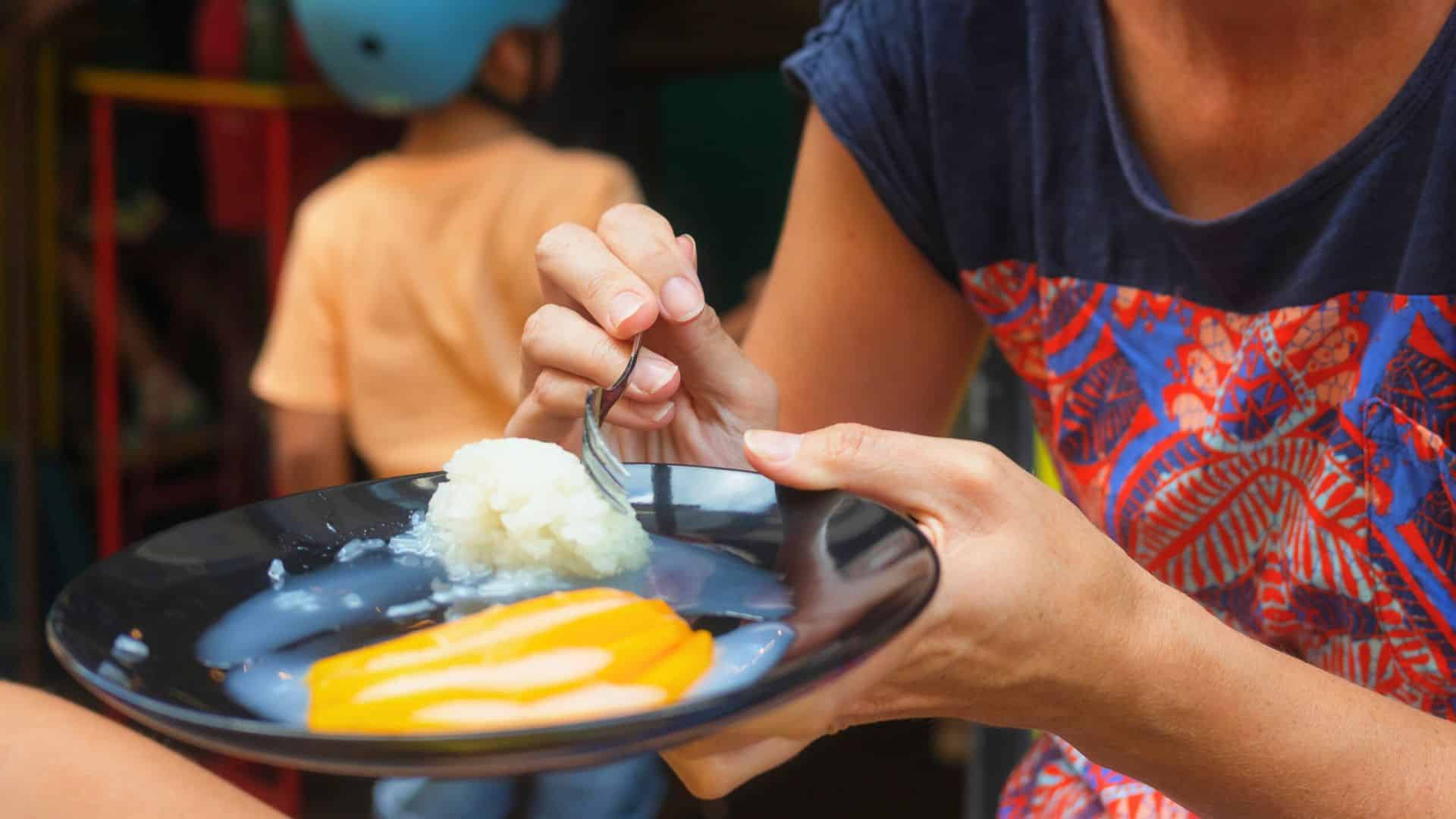 A person holding a plate of mango sticky rice, about to eat with a fork; child blurred in the background.
