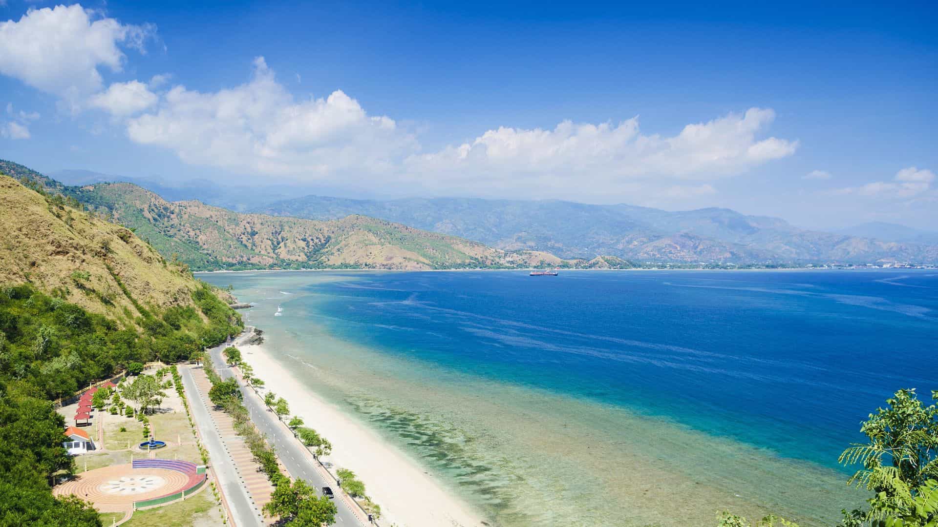 A curving coastline with clear blue water, sandy beach, green hills, and mountains under a bright blue sky.