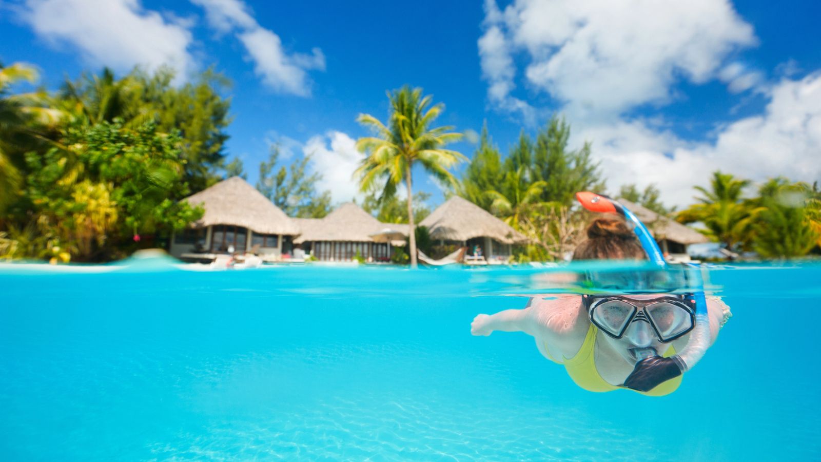Person snorkeling in clear blue water near tropical huts and palm trees under a bright, sunny sky.