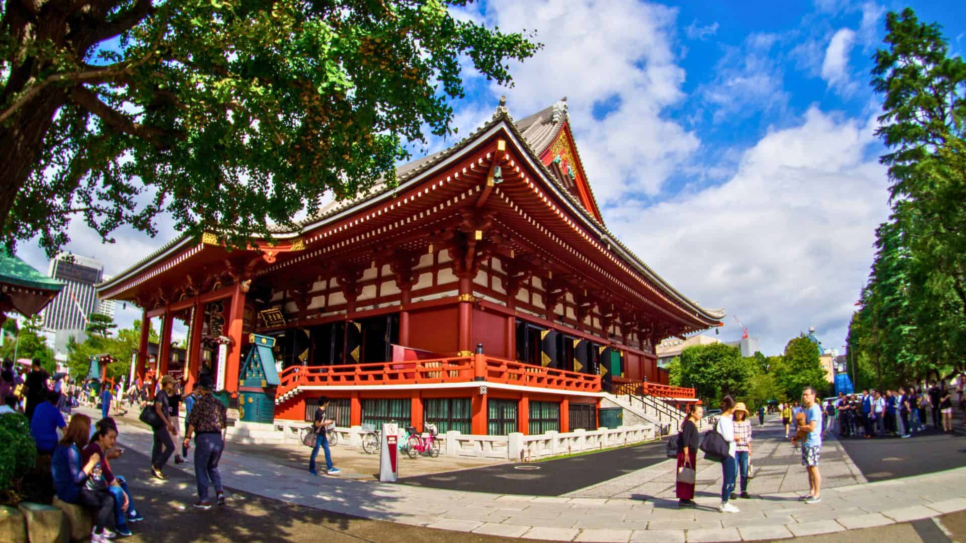 People walking near a large traditional Japanese temple with a red exterior and sloped roof on a sunny day.