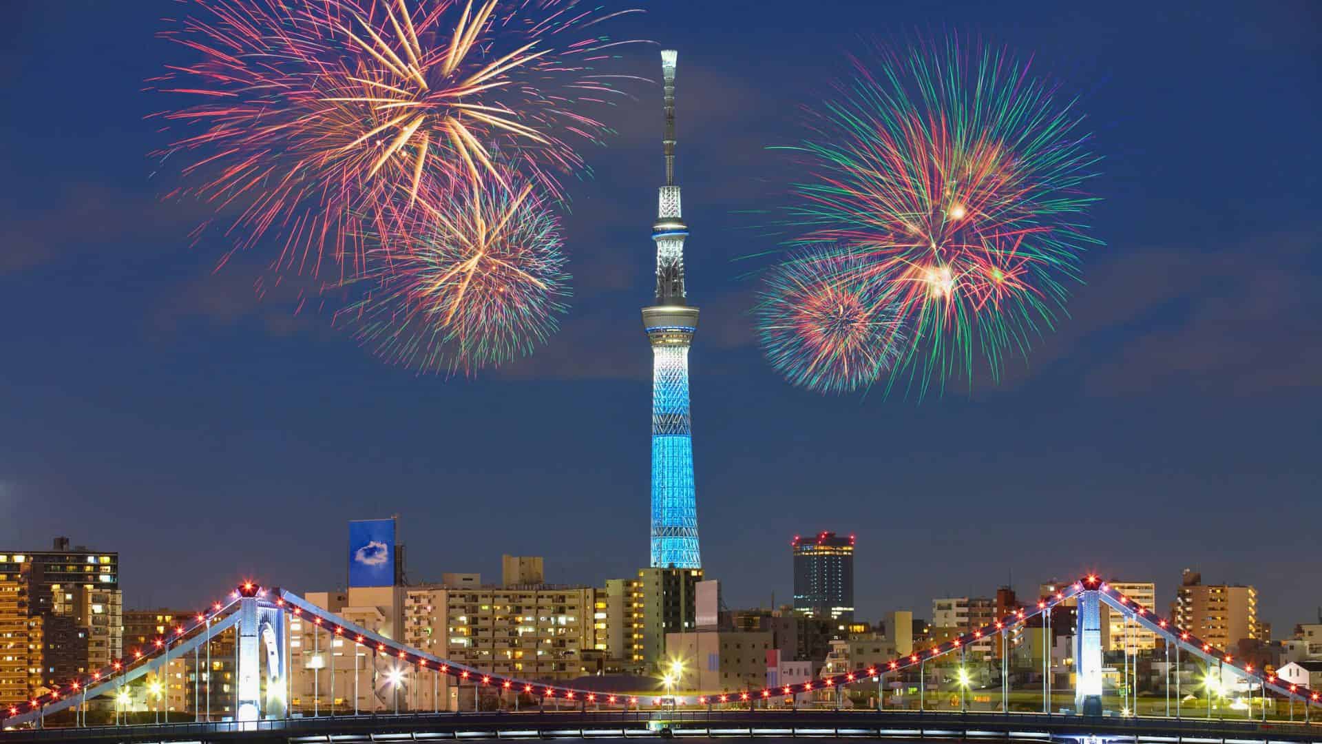 Fireworks light up the night sky above Tokyo Skytree and city buildings, with a lit-up bridge in the foreground.