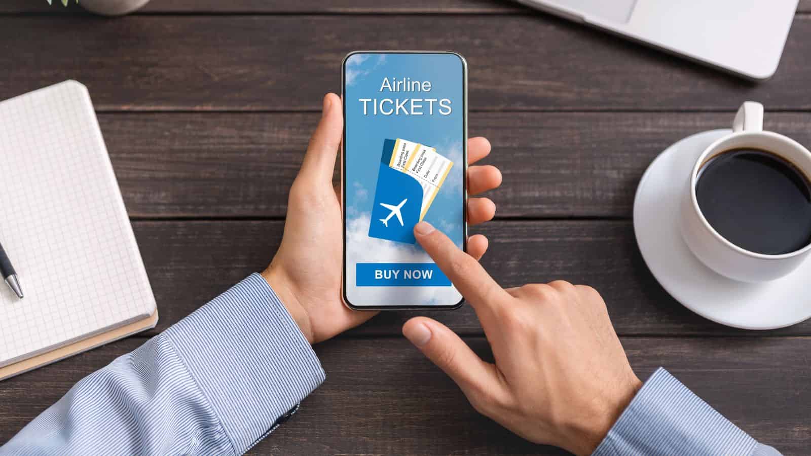Person holding a phone with an airline tickets booking app, next to a notebook, laptop, and coffee cup.