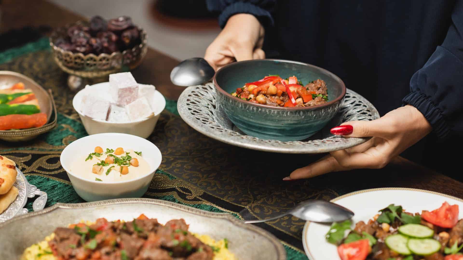 A person serves a bowl of stew among dishes of salad, rice, and sweets on a decorated table.