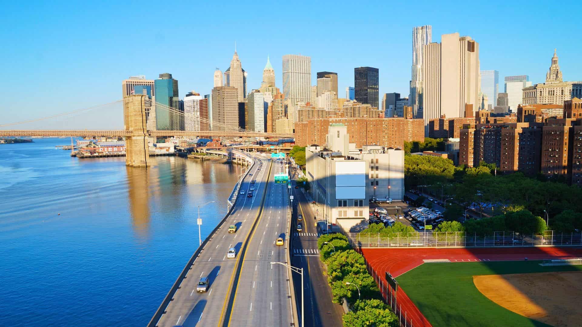 A sunny view of Manhattan skyline, Brooklyn Bridge, and cars driving along the waterfront highway.