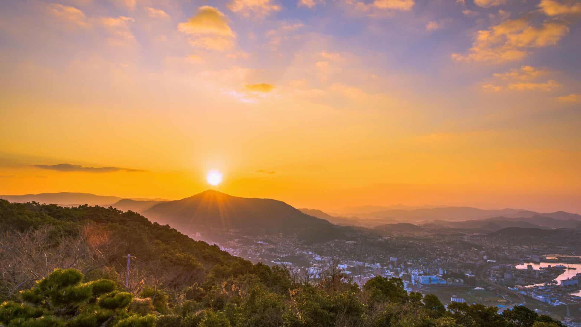 Sunrise over a city with mountains in the background, trees in the foreground, and a colorful sky.